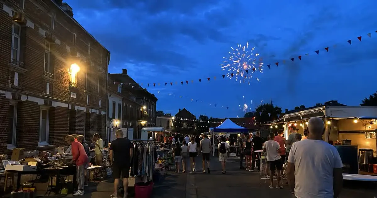 Fête de village en soirée : brocante dans la rue, guirlandes de fanions tricolores, stands et feu d'artifice dans le ciel bleu nuit