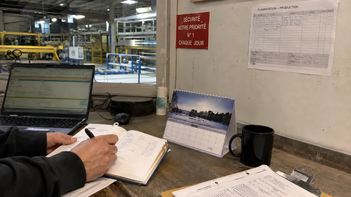 Calendrier chevalet de bureau posé dans un atelier industriel, près d'un planning de production et d'un agenda ouvert